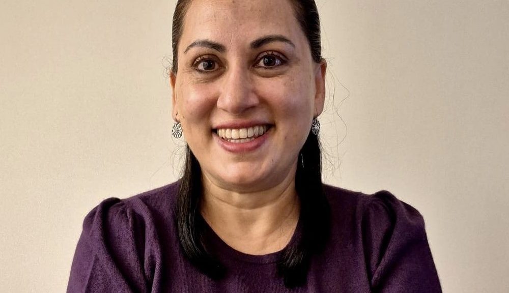 A smiling woman with dark hair pulled back, wearing silver earrings and a purple top, poses against a plain beige background.