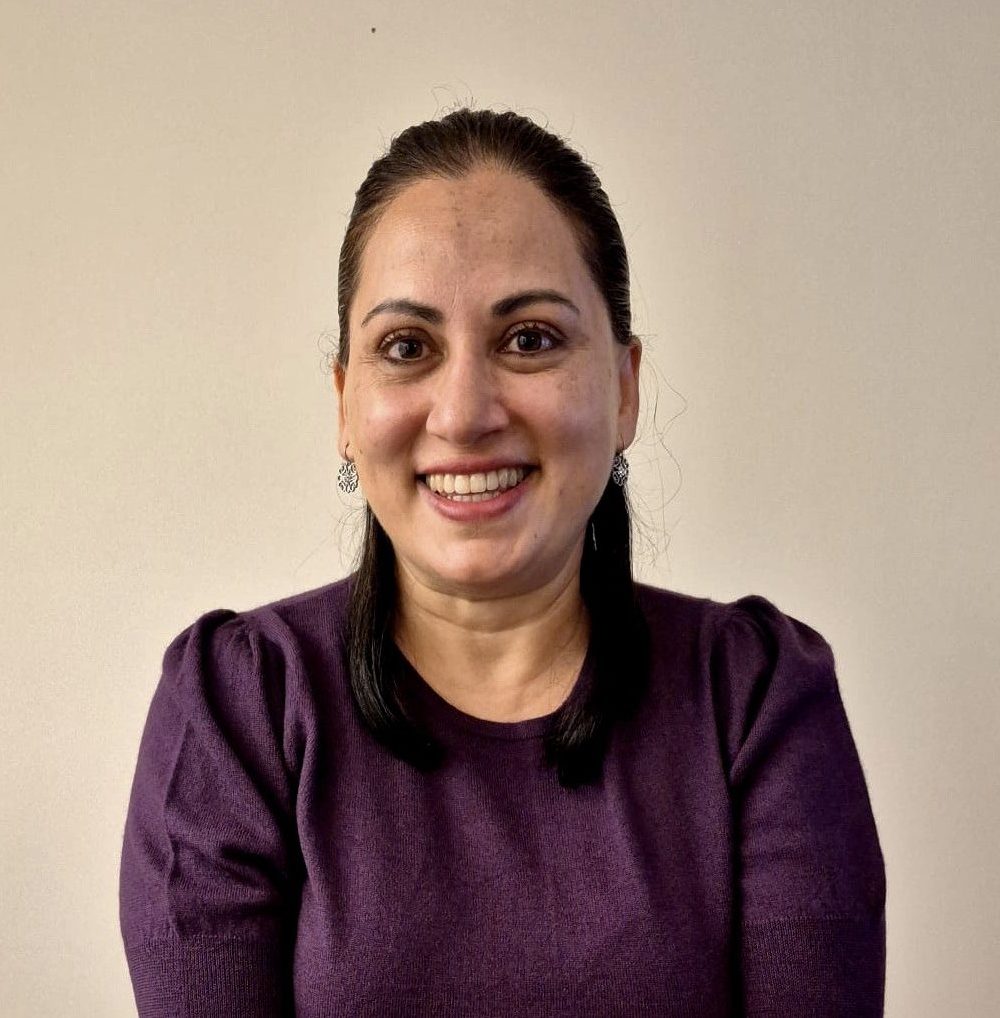 Leah SK photo A smiling woman with dark hair pulled back, wearing silver earrings and a purple top, poses against a plain beige background.