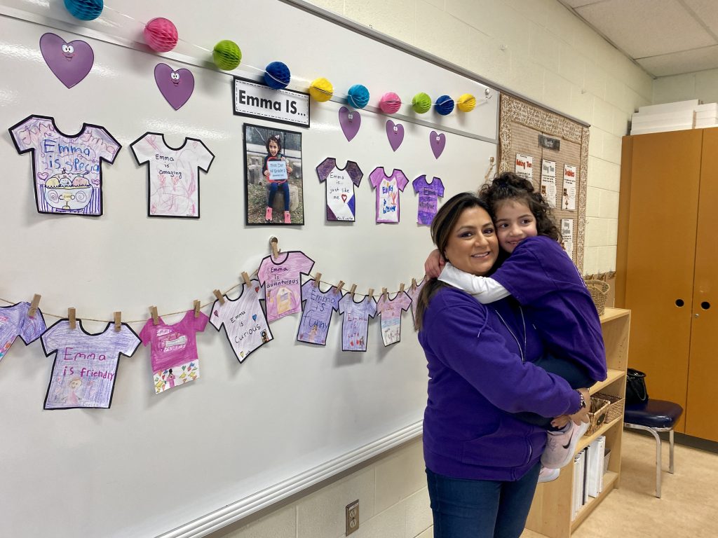 Monica holds Emma, both in purple shirts, standing in a front of a classroom white board covered in children's art work of cut out t-shirts.