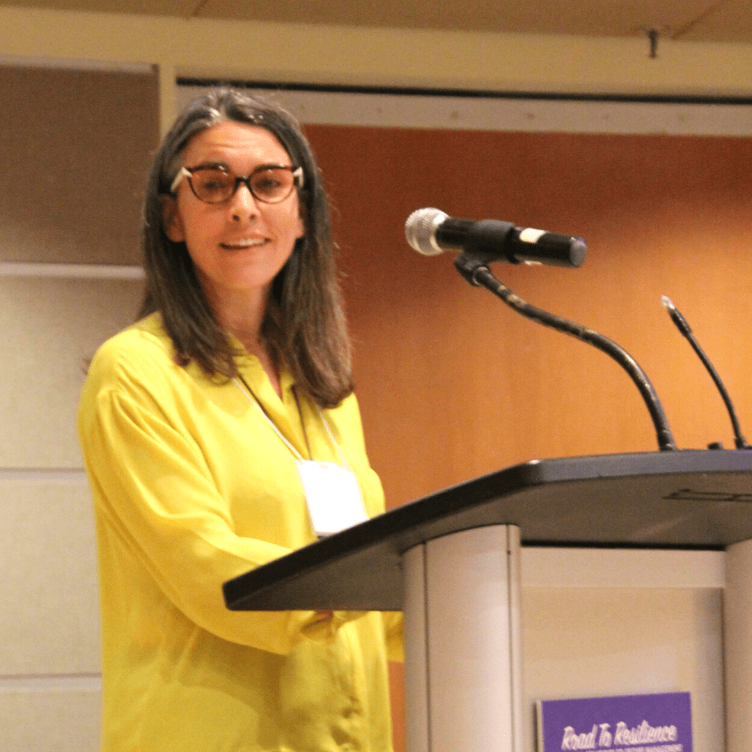 A woman wearing glasses and a yellow blouse stands behind a podium