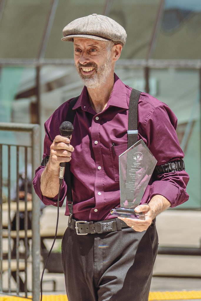 Max T Oz, wearing a purple button up shirt, suspenders and a newsboy cap, stands outside holding a glass HOPE award and microphone.