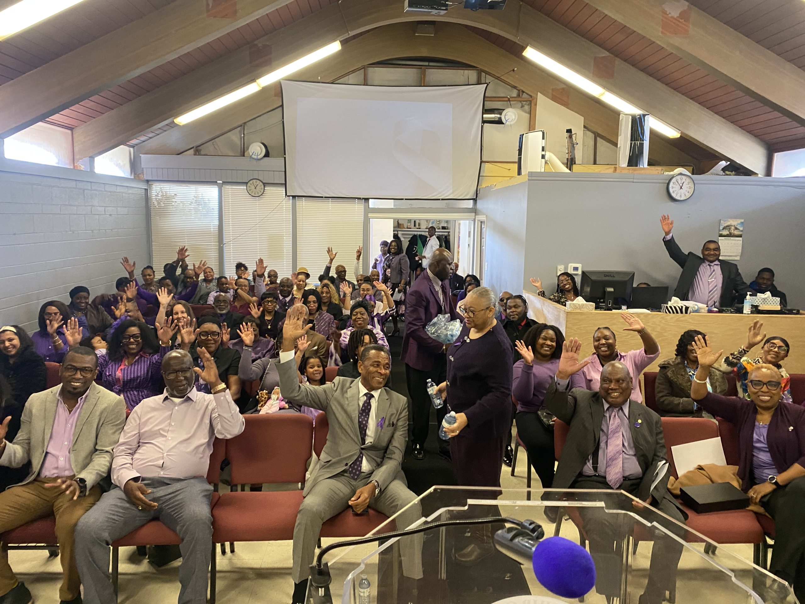 A congregation of people, many wearing purple, smile and wave.