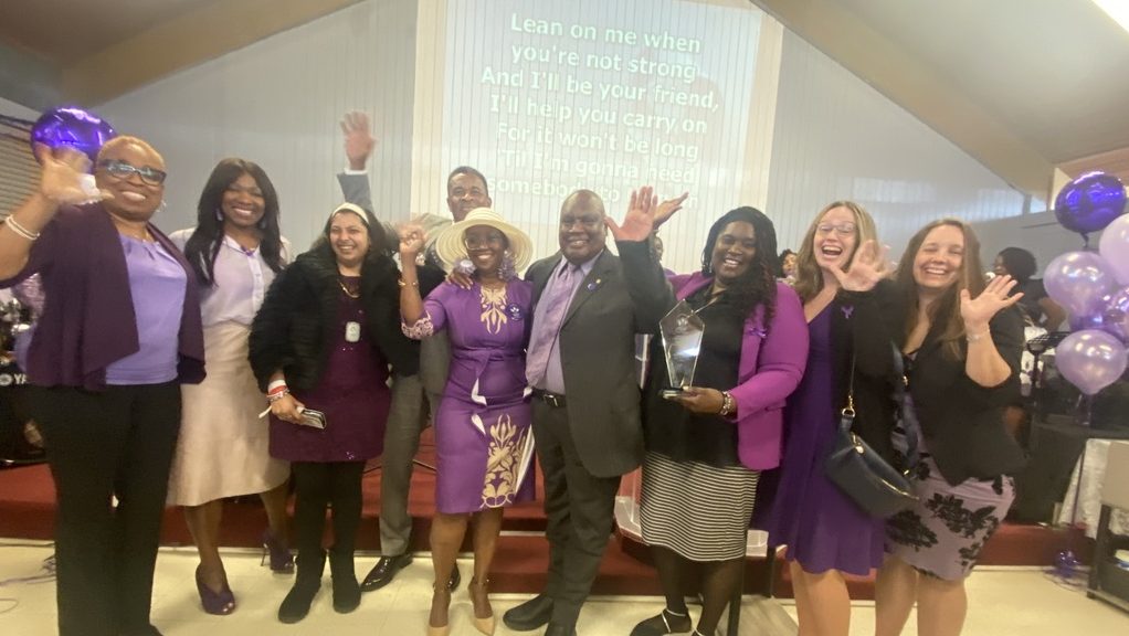 A group of diverse people dressed in suits and dresses stand smiling and waving.