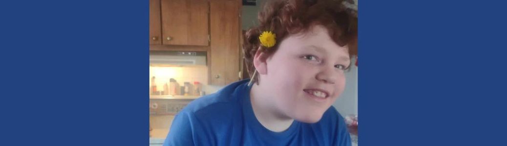 Landyn smiles at camera with a dandelion behind his ear.