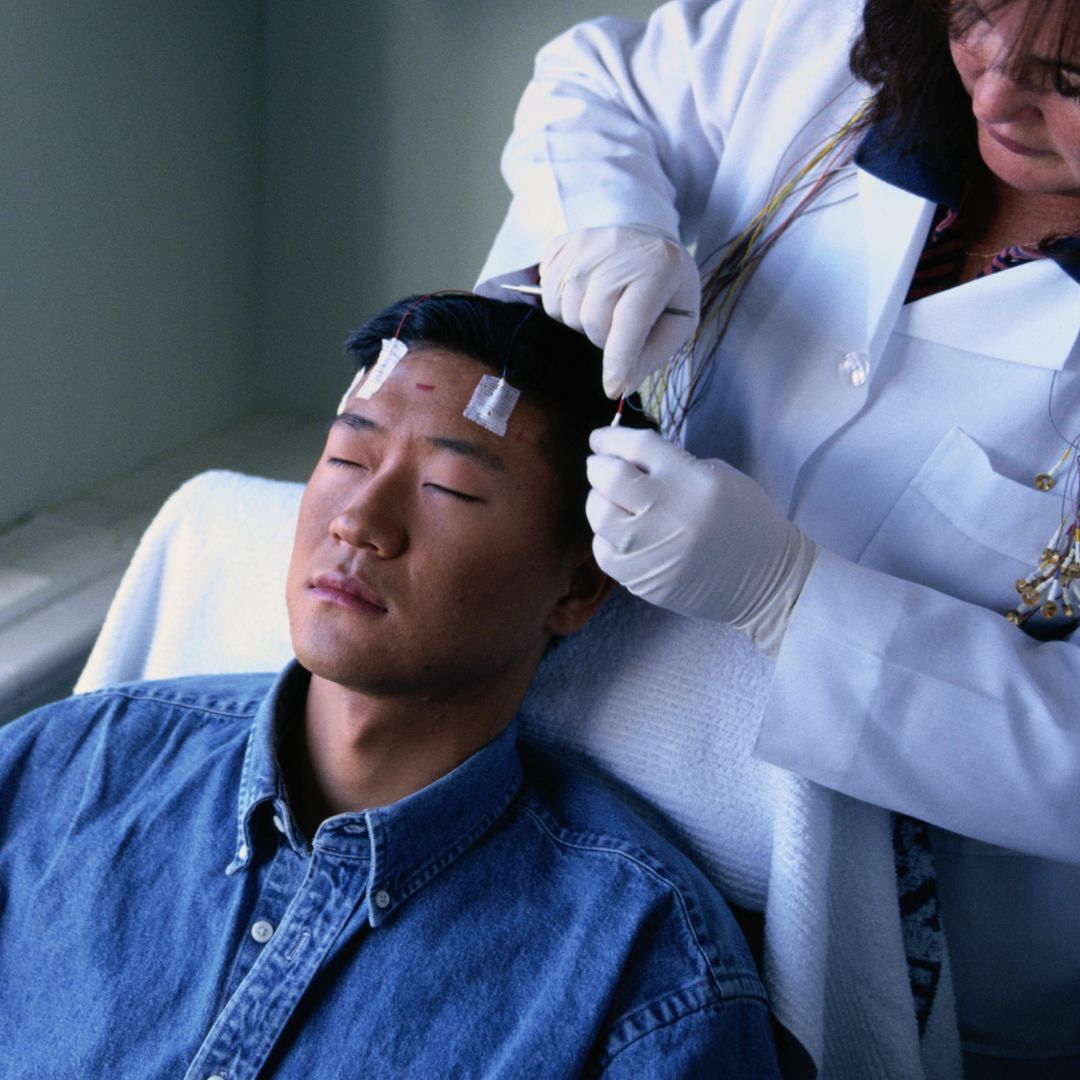 Man sits back on a chair, with his eyes closed as a doctor affixes electrodes to his head.