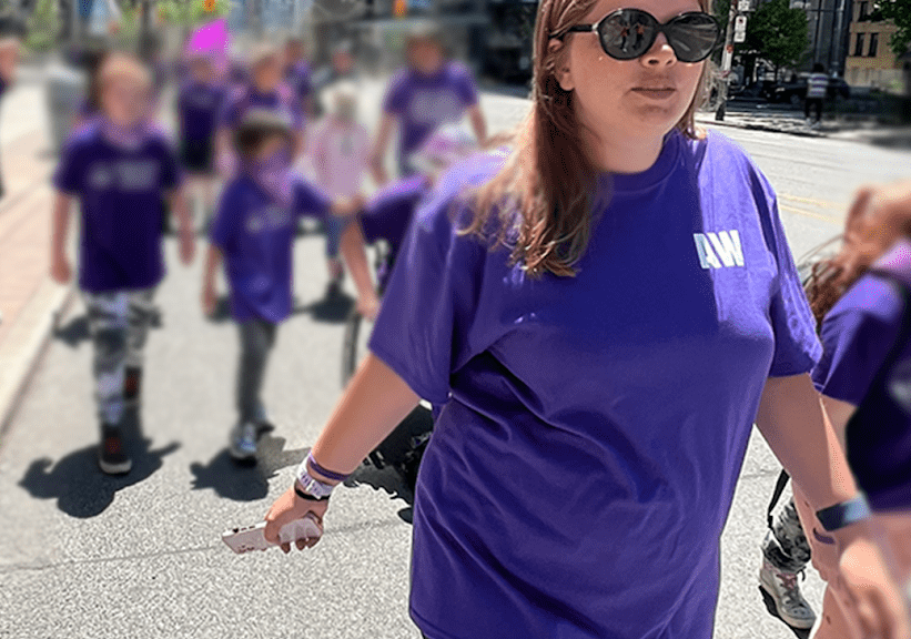 Woman in a purple t-shirt and sunglasses walks proudly down the centre of the street with other people dressed in purple blurred behind her.