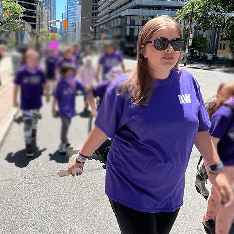 Woman in a purple t-shirt and sunglasses walks proudly down the centre of the street with other people dressed in purple blurred behind her.