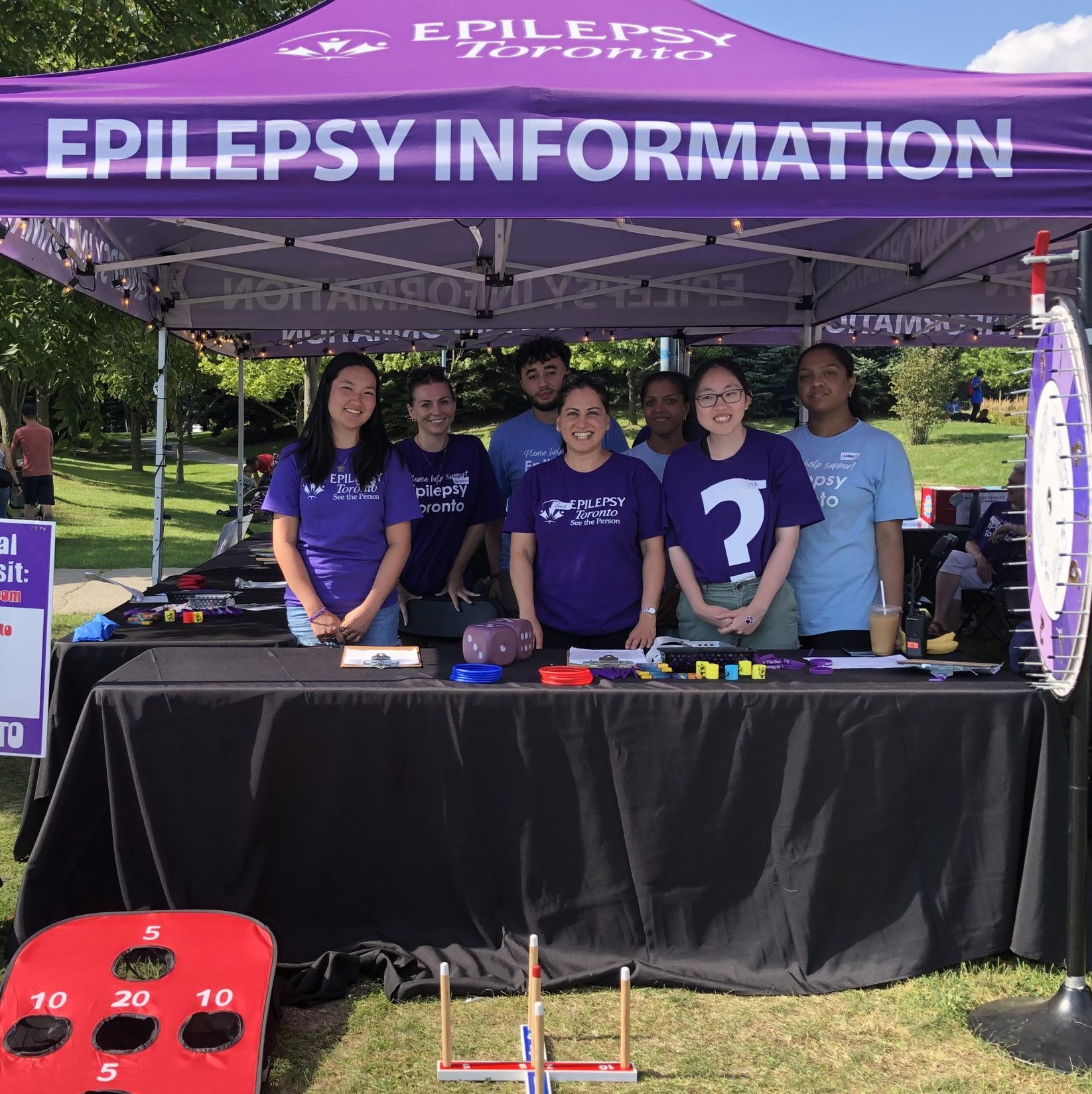 A diverse group of smiling people stand under a purple Epilepsy Information tent at the Toronto International BuskerFest for Epilepsy.