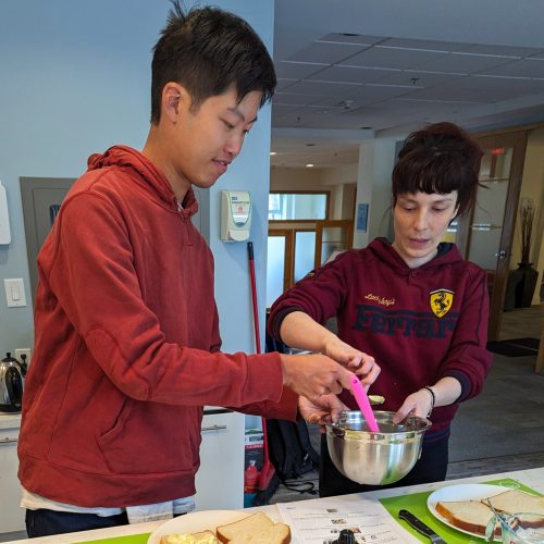 Two people stand at a kitchen counter making sandwiches. One person in a red hoodie spreads egg salad onto bread while the other holds a mixing bowl. Cooking utensils, mustard, lettuce, and a carrot are on the counter.