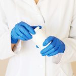 The torso of a person in a white lab coat, wearing blue rubber gloves and opening an medication bottle.
