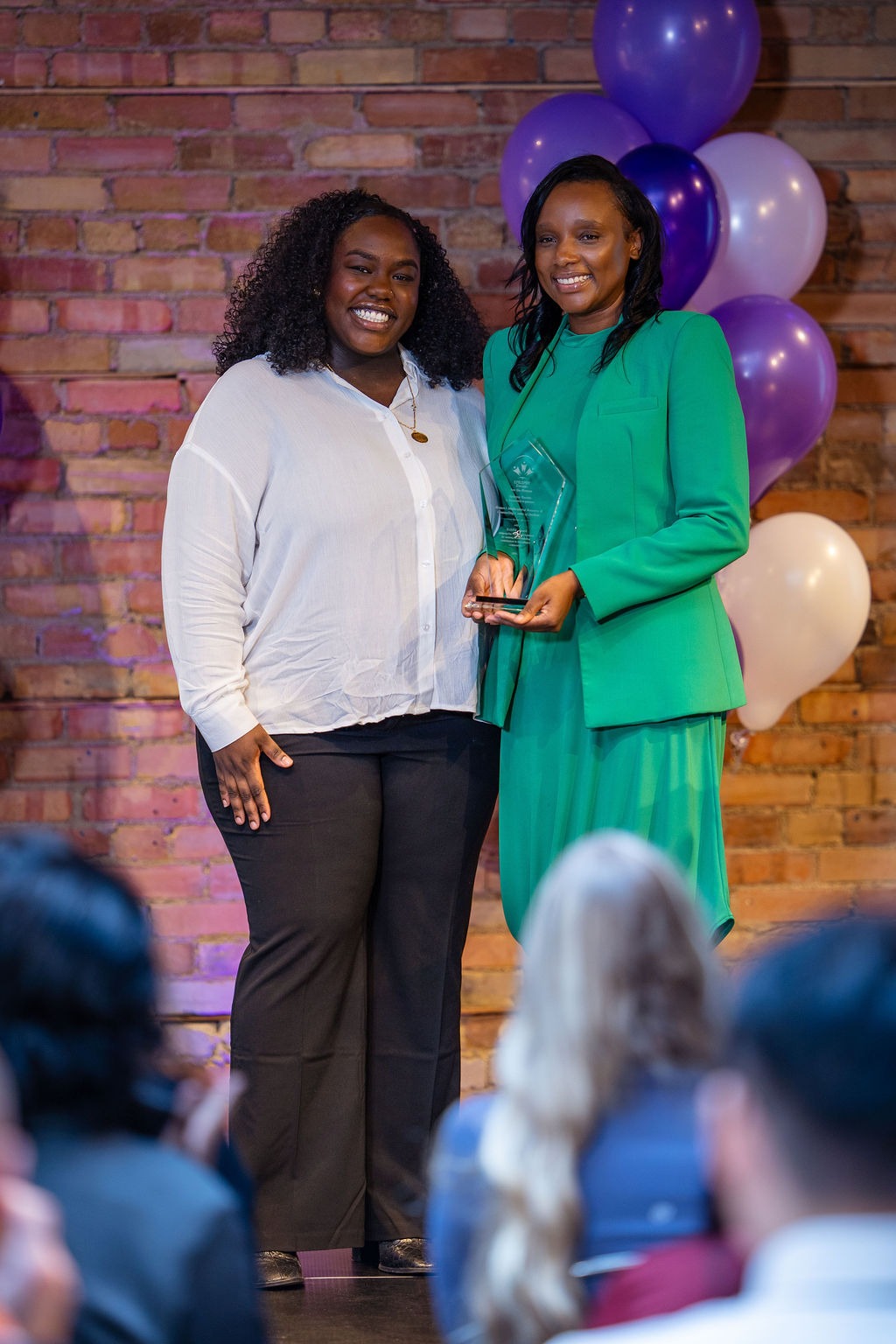 Two smiling women pose with a glass award at an Epilepsy Toronto SeizeABILITY! recognition event. One wears a white blouse, while the other wears a bright green suit and holds the award. Purple and white balloons decorate the brick wall behind them, with audience members seated in the foreground.